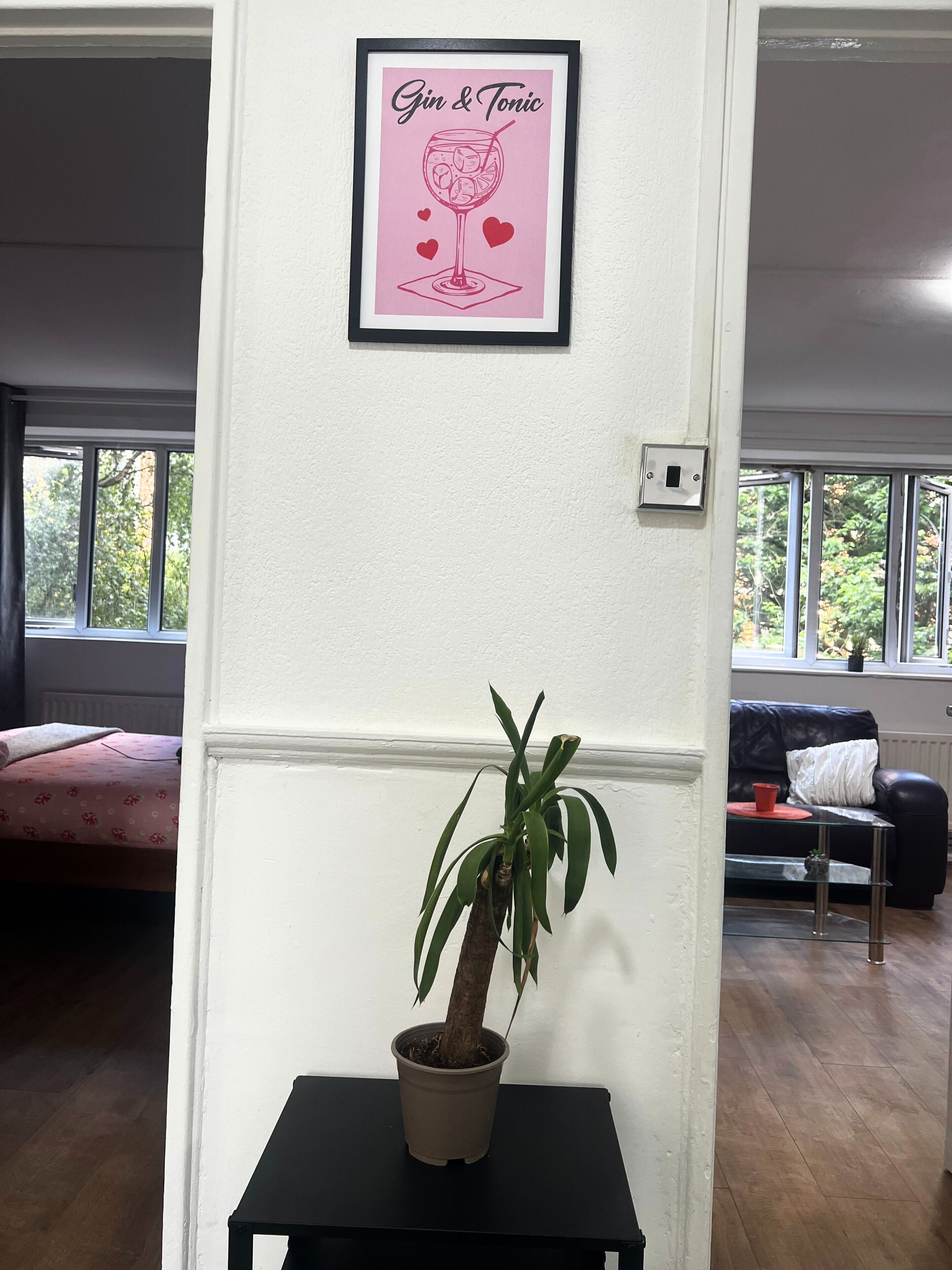 Quarto com quadro de um gin tônico na parede, planta em vaso sobre uma pequena mesa, um sofá e janela com vista para árvores.