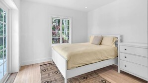 Een slaapkamer met een bed, nachtkastje, dressoir en een raam met uitzicht op groen.