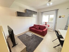 Un espace de vie compact comprenant un canapé rouge, une petite table à manger, un téléviseur et une fenêtre avec des rideaux.
