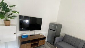 Una sala moderna con un televisor de pantalla plana montado en la pared, un sofá gris, un mueble de entretenimiento de madera, un refrigerador y una planta en maceta.