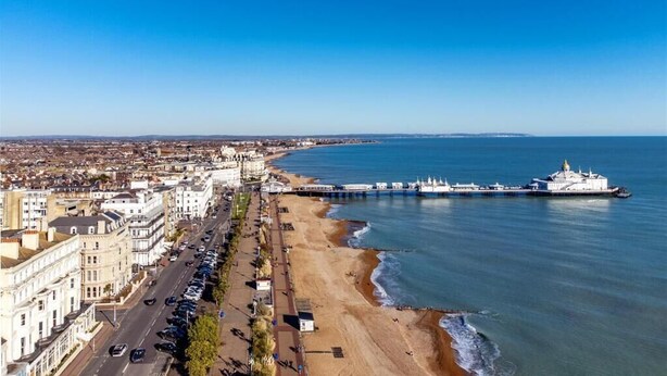 Un paisaje urbano costero con un muelle largo que se extiende hacia el mar, bordeado de edificios y una carretera paralela a la playa.
