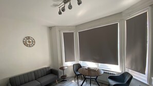 Un moderno salón con un sofá gris, una mesa redonda y dos sillas. Se observan ventanas amplias con persianas, un ventilador de techo y un reloj de pared.