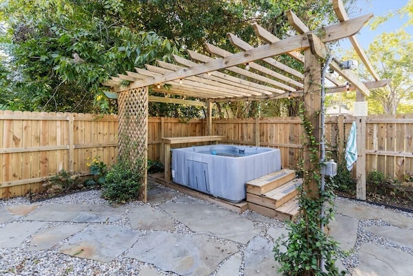 A wooden pergola structure with a hot tub underneath, surrounded by a wooden fence and stone pathway.