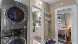 A laundry room with a front-loading washer and dryer, a bathroom with a vanity and mirror, and a bedroom with a bed and window. - Jasmine Cottage - Hot Tub - Very Pet Friendly (Wilmington)