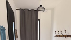 Un couloir avec une porte sombre, un rideau gris, une chaise en osier, une décoration murale en macramé et un vase vert posé sur le sol.