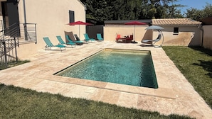 Une piscine entourée de chaises longues et de parasols, elle-même bordée d’une terrasse carrelée et d’une pelouse.