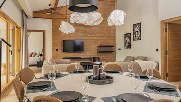 A modern dining area with a round table set for six, surrounded by chairs. A television is mounted on the wooden wall, and there are two large, white, crumpled paper lampshades hanging above the table.