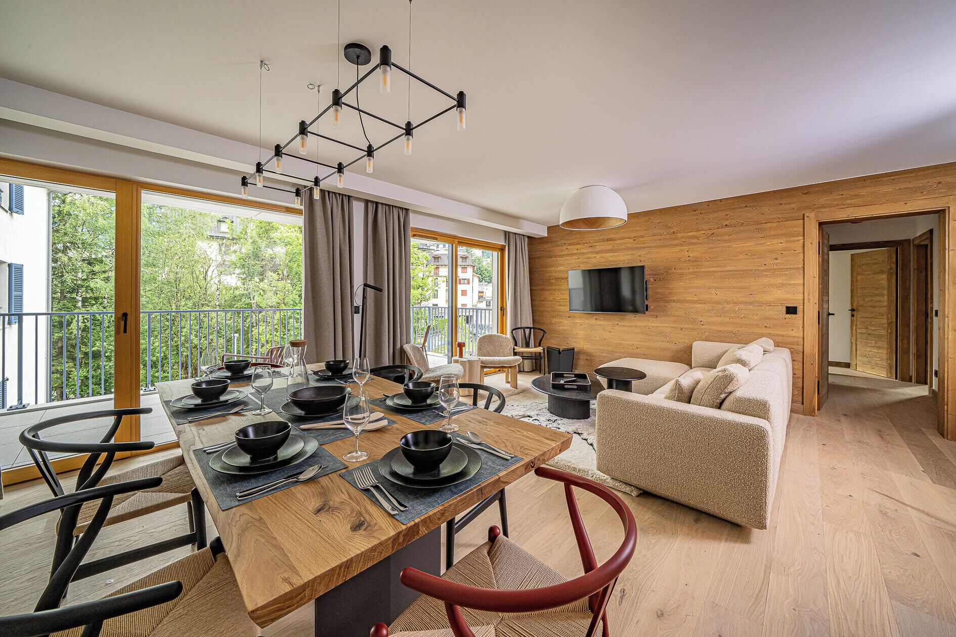 Ein modernes Wohnzimmer mit Essbereich, Holzvertäfelung, einem an der Wand montierten Fernseher und einem Balkon mit Blick auf Grünflächen.
