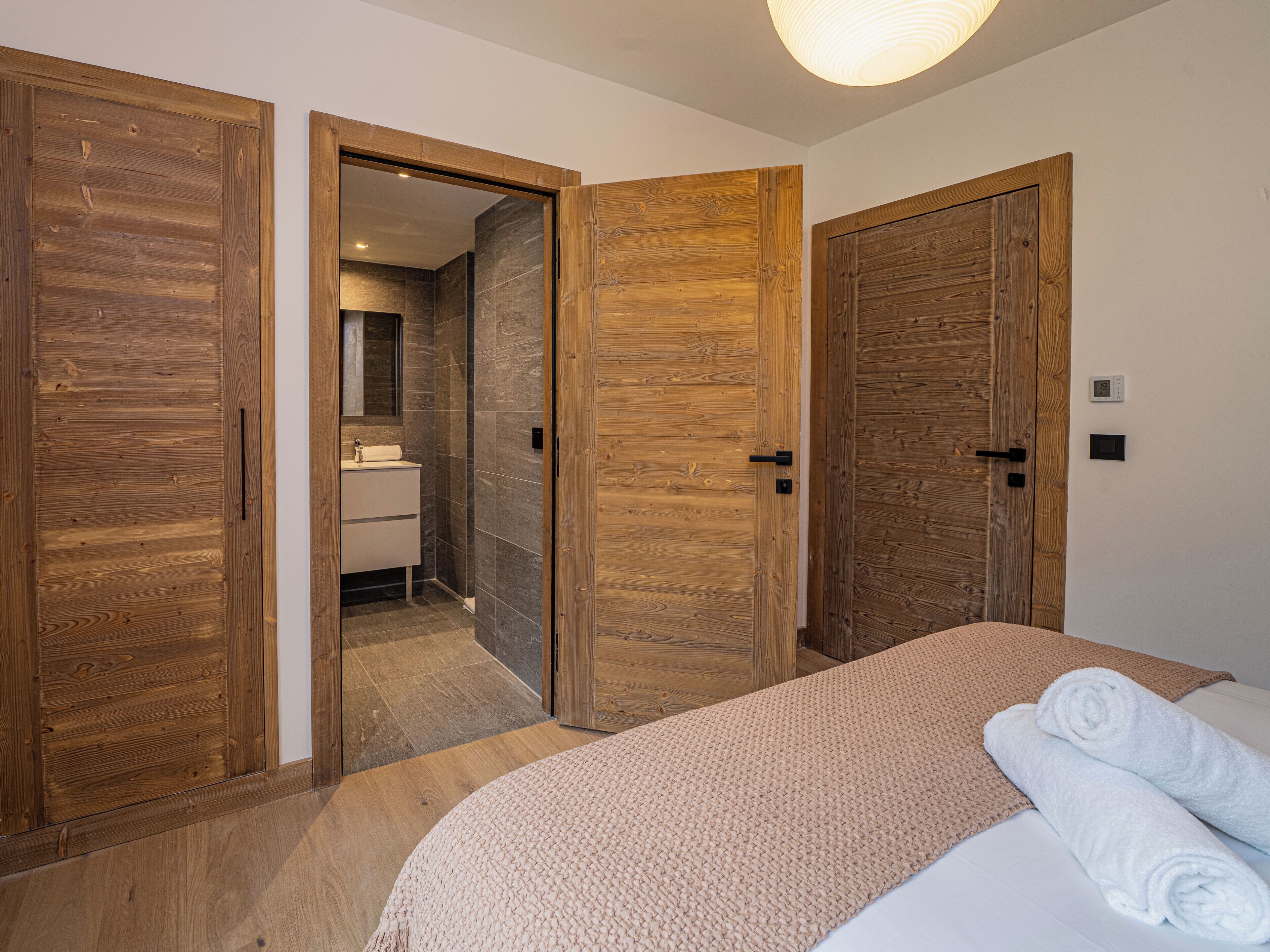Une chambre avec des portes en bois, un lit recouvert d’une couverture beige et une salle de bain visible à travers une porte ouverte.
