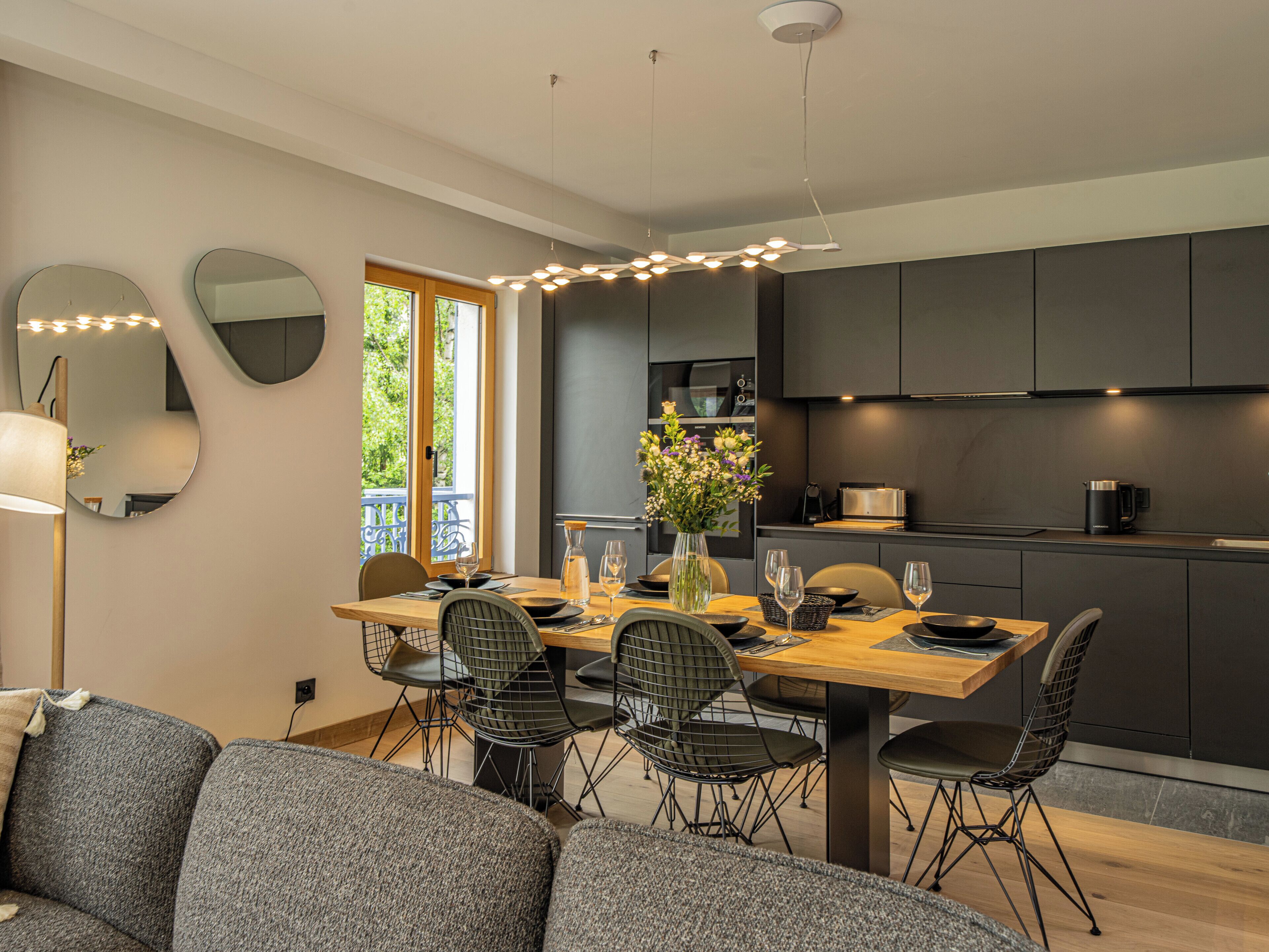 Ein moderner Essbereich mit Holztisch, schwarzen Stuhlgestellen und einem Blumenstrauß. Im Hintergrund ist eine Küche mit dunklen Schränken und einer Kaffeemaschine zu sehen.