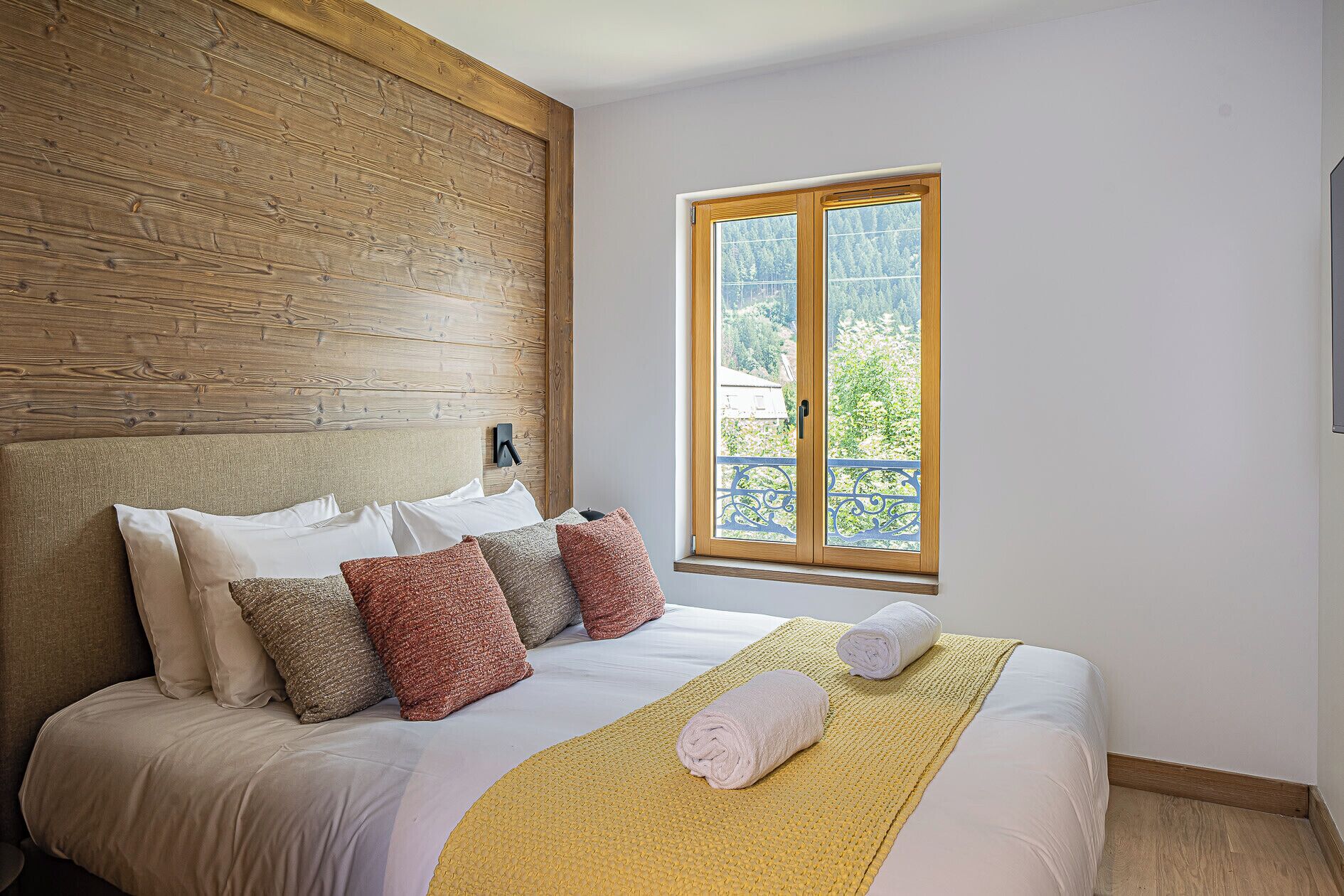 A bedroom with a wooden headboard, a bed with white bedding and multiple pillows, a yellow throw blanket, and a window with a view of greenery and buildings.