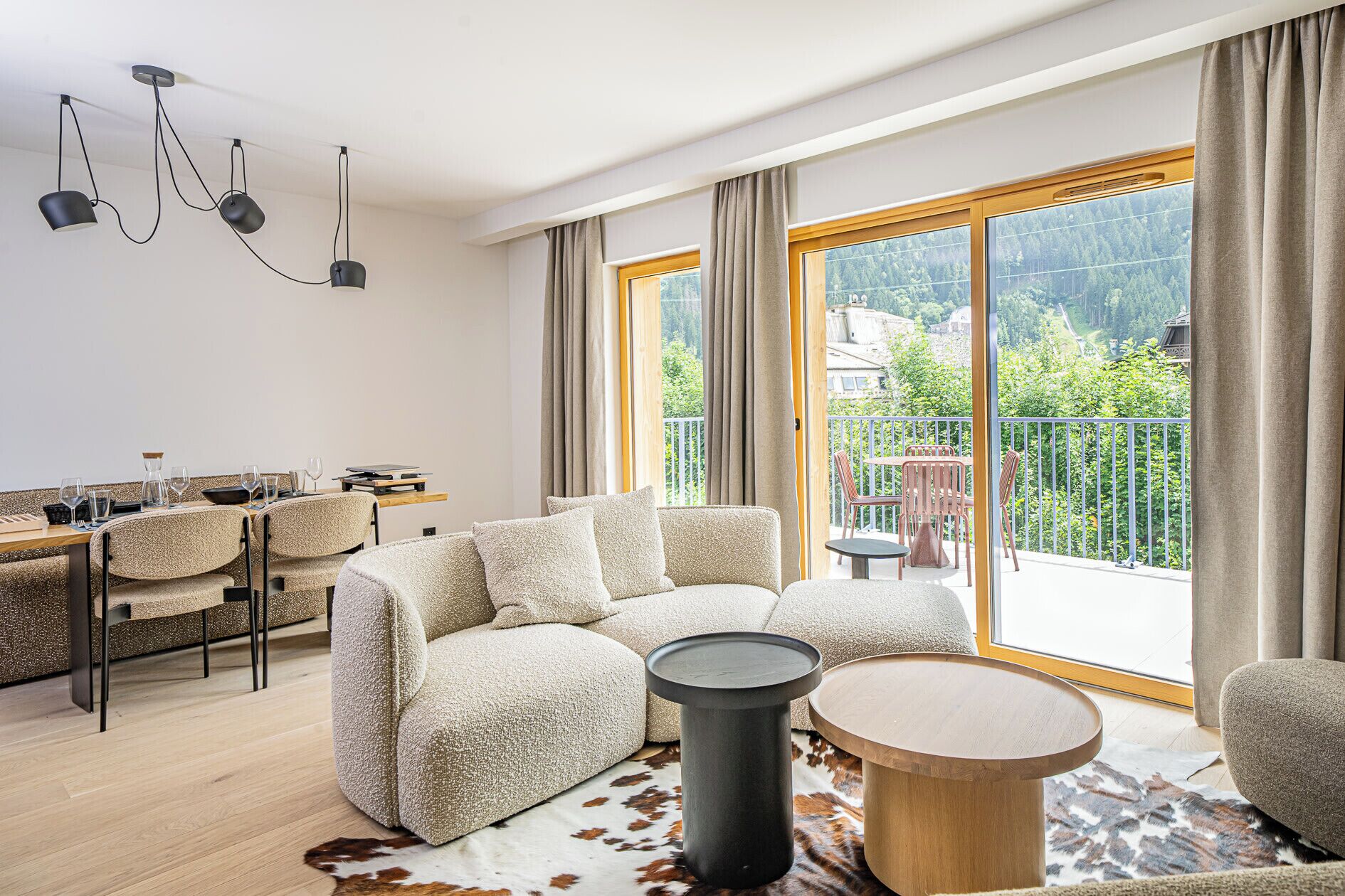 A modern living room with a sofa, a coffee table, and a dining area with chairs and a table. There is a balcony with a view of greenery and buildings.