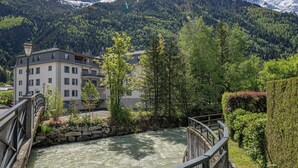 Een hotel aan de rivier met een stenen brug, omgeven door weelderige begroeiing en bergen op de achtergrond.
