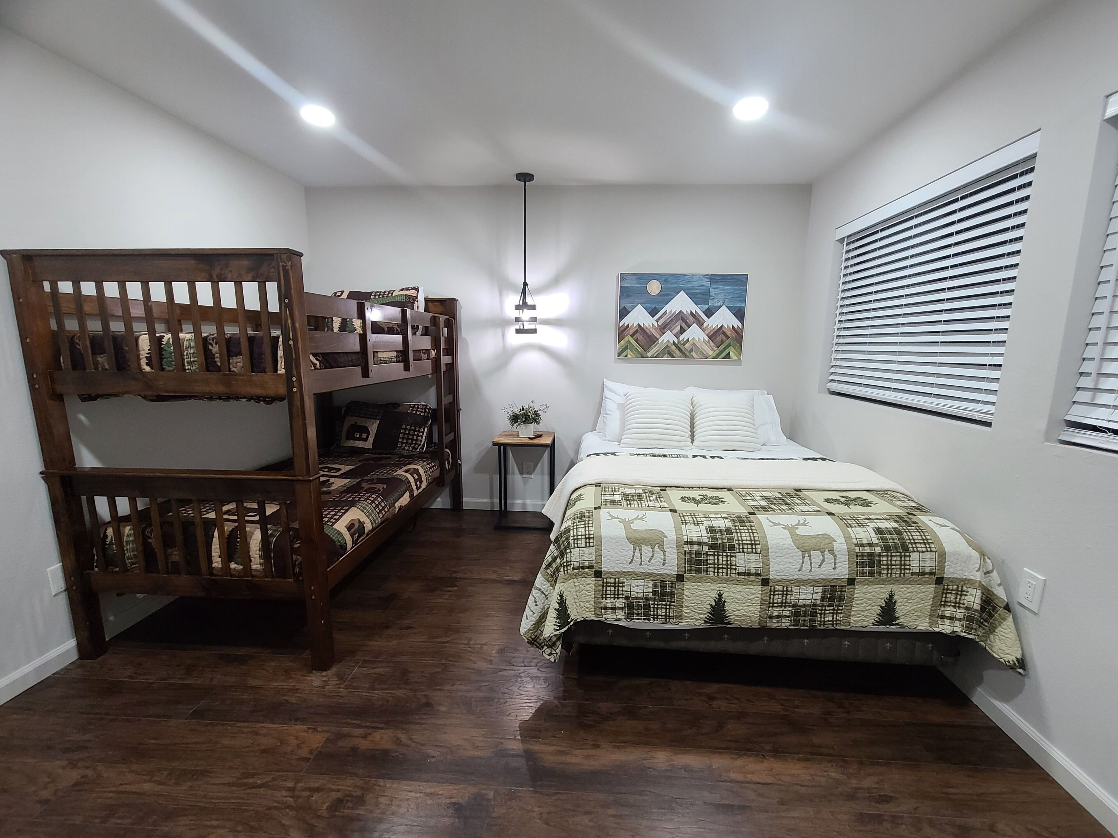 A room with a bunk bed, a single bed with a quilt, a small table with a plant, a wall-mounted picture, and a window with blinds.