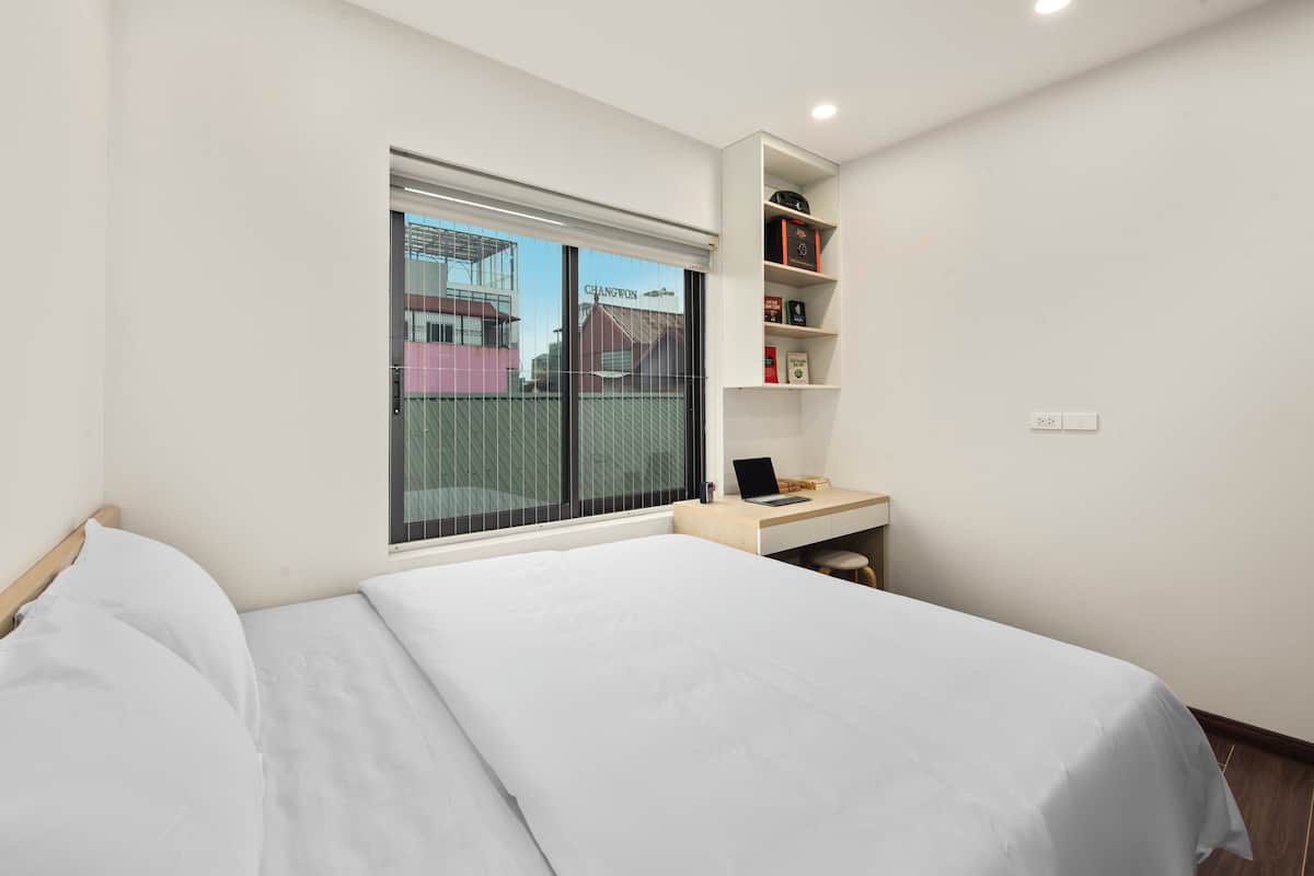 Quarto moderno com cama grande, escrivaninha com laptop e janela com vista para edifícios.