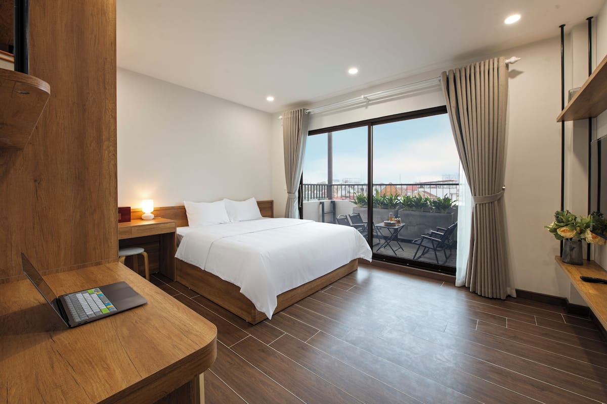 Quarto de hotel moderno com uma cama grande, uma escrivaninha de madeira, um laptop, uma varanda com vista e uma TV sobre uma estante de madeira.