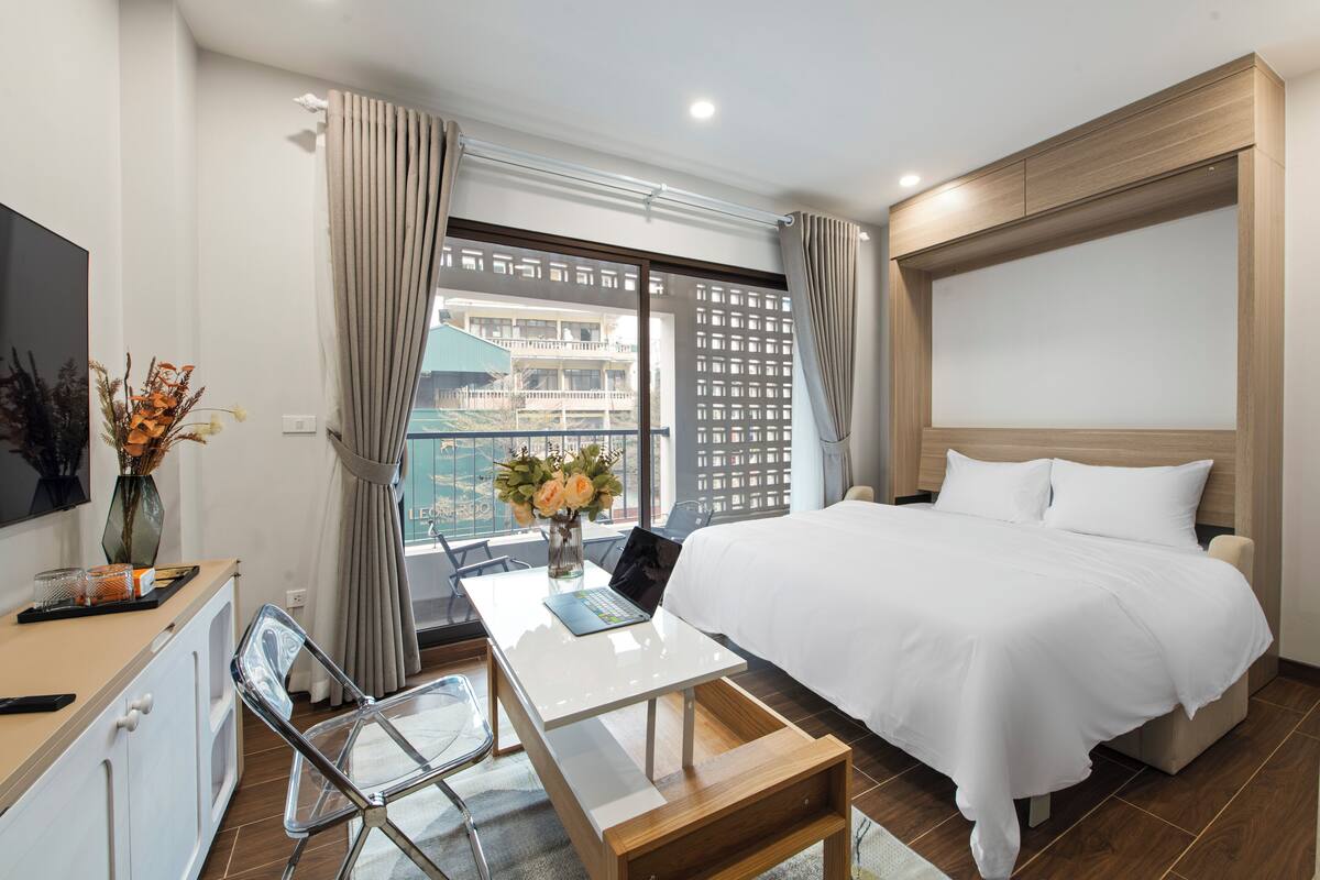 Quarto de hotel moderno com cama, escrivaninha com laptop, televisão e vista para um edifício pela janela.