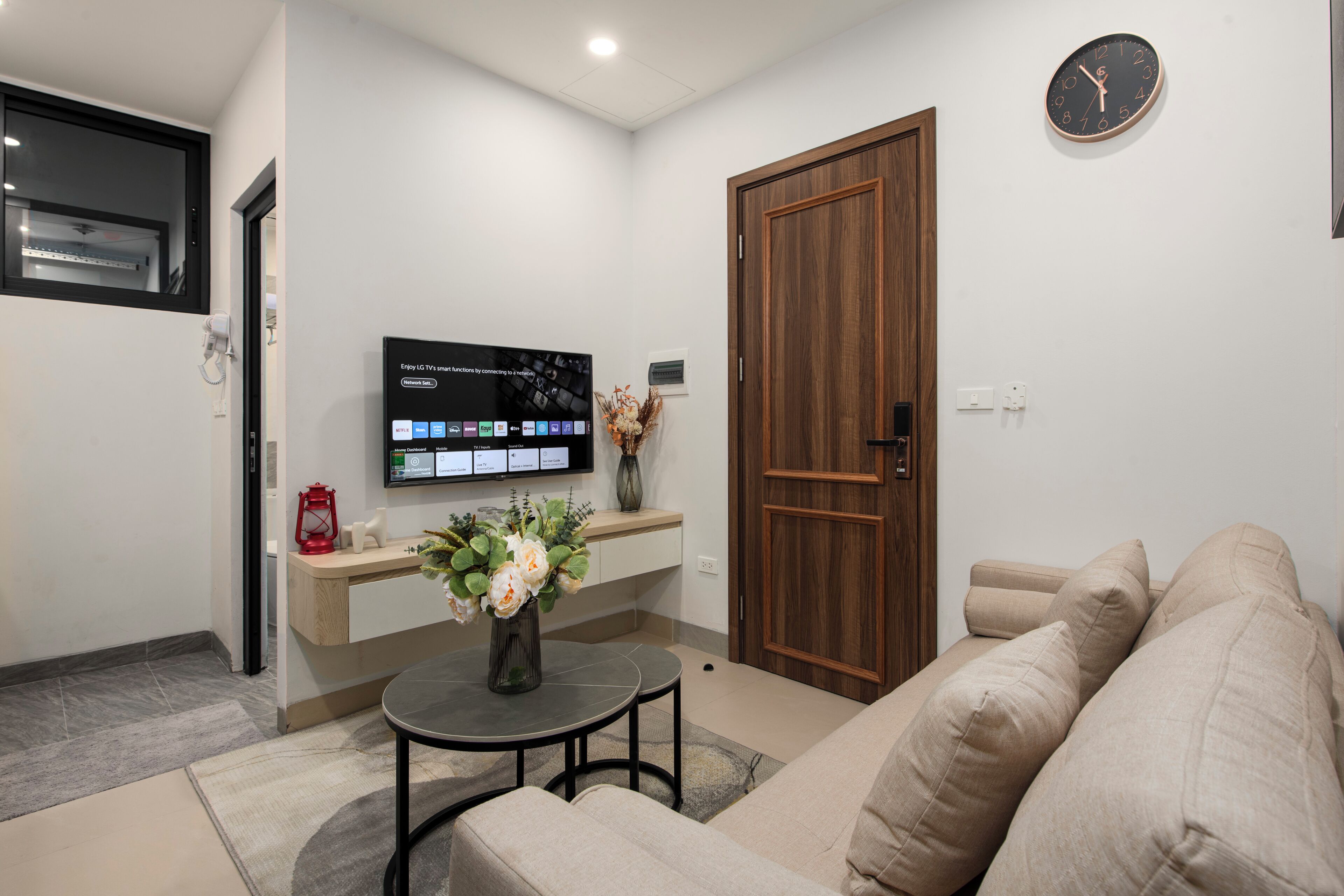 A modern living room with a flat-screen TV mounted on the wall, a wooden door, a beige sofa, and a small round table with a vase of flowers.