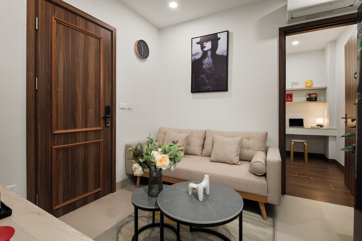 Uma sala de estar com uma porta de madeira, um sofá, uma mesa de centro, um vaso com flores e uma moldura com uma fotografia na parede.