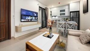 Un salon moderne doté d’un téléviseur à écran plat, d’un coin repas avec une table et des chaises blanches, et d’une cuisine avec des meubles blancs et des appareils en acier inoxydable.