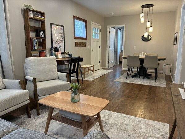 Un salon comprenant un canapé, une table basse, une bibliothèque, un coin repas avec des chaises, et une cuisine à l’arrière-plan.