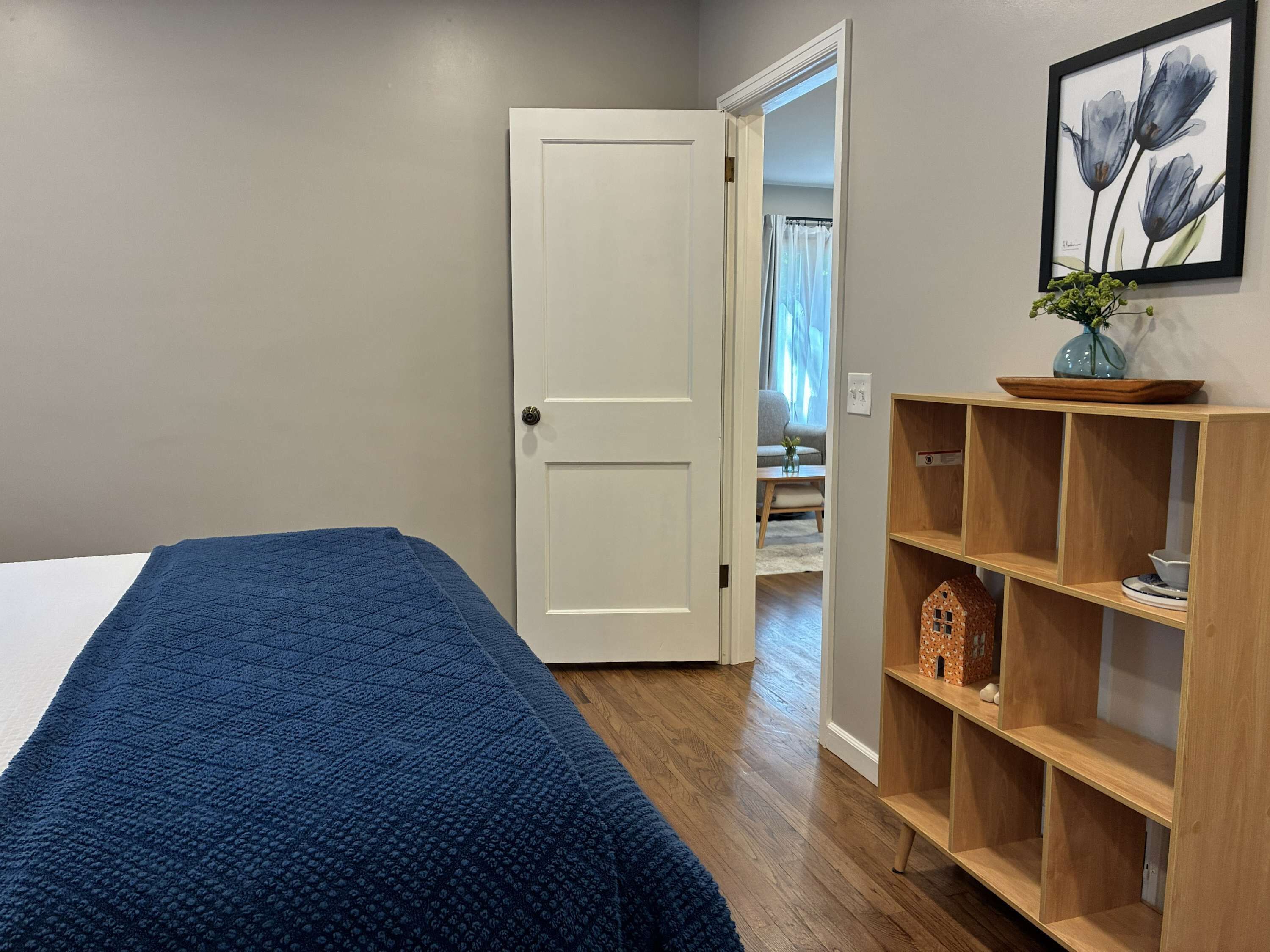 Ein Schlafzimmer mit einem Bett, einer Tür, einem Holzregal mit Dekoration und einem gerahmten Bild an der Wand.