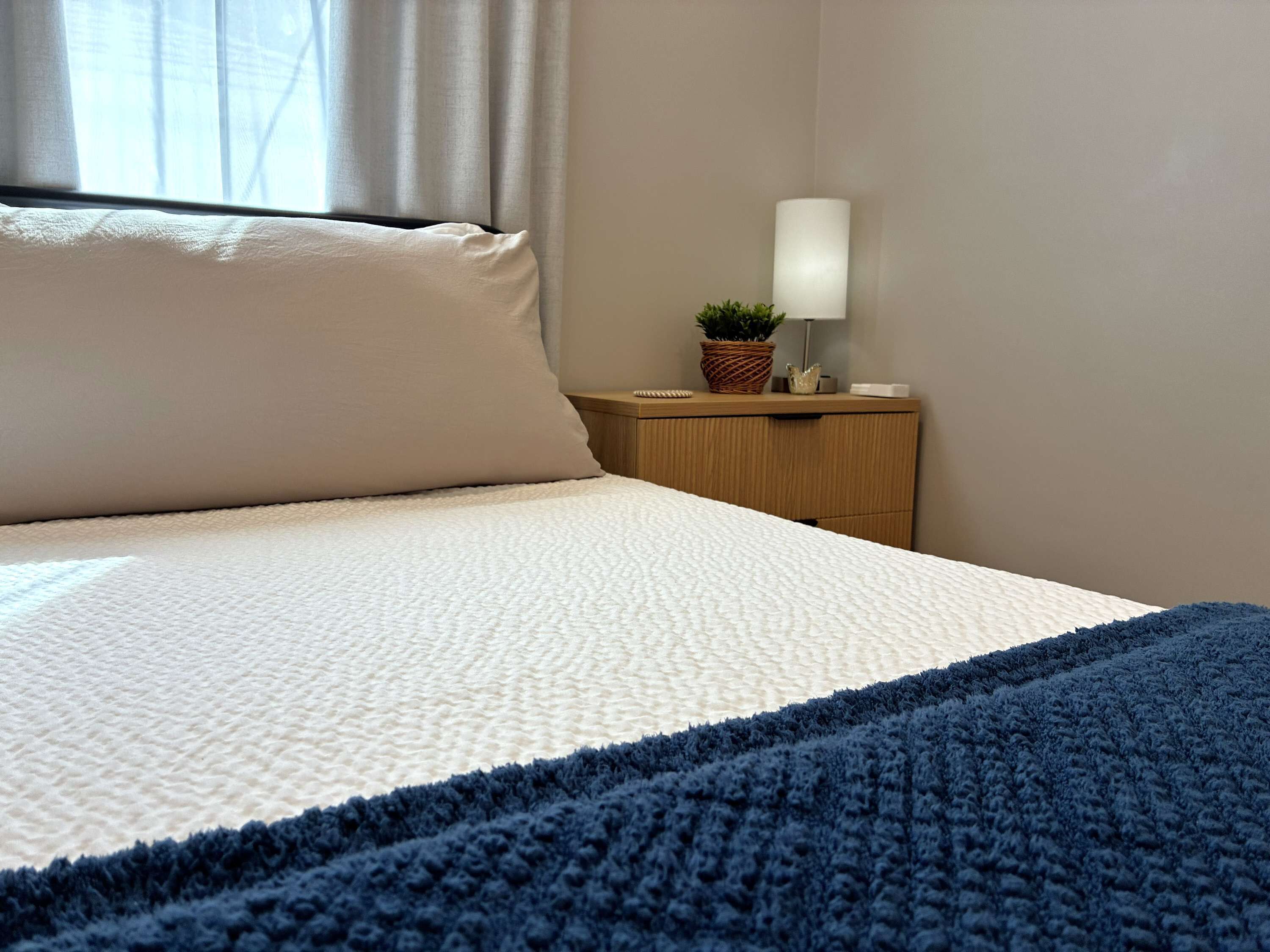 A neatly made bed with a textured white cover and a blue blanket, a bedside lamp, and a small potted plant on a wooden nightstand.