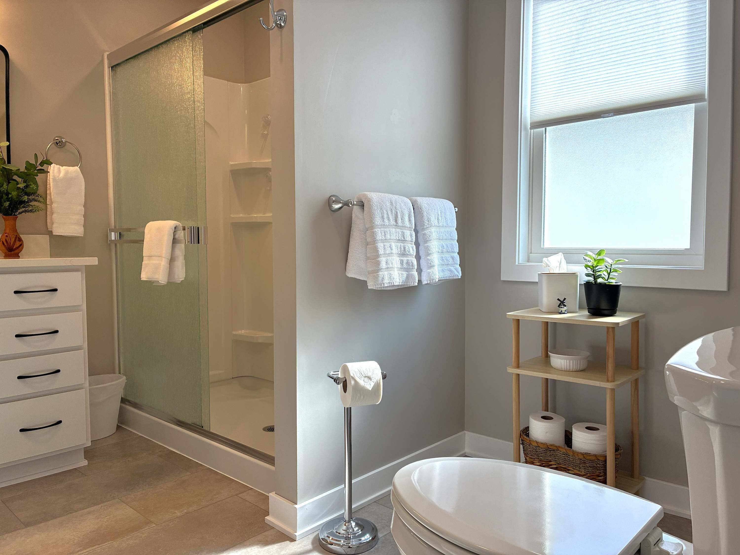 A modern bathroom with a glass-enclosed shower, white fixtures, and a window providing natural light.