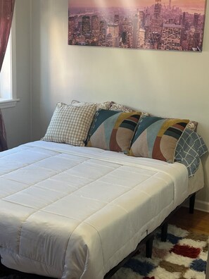 A neatly made bed with a white duvet, a cityscape painting on the wall, and a patterned rug on the floor.