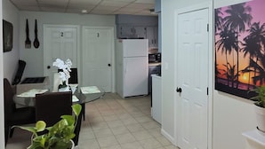 Una cocina compacta con armarios blancos, un refrigerador y un microondas. Hay un área de comedor con una mesa de vidrio y sillas, y una planta en maceta sobre una mesita.