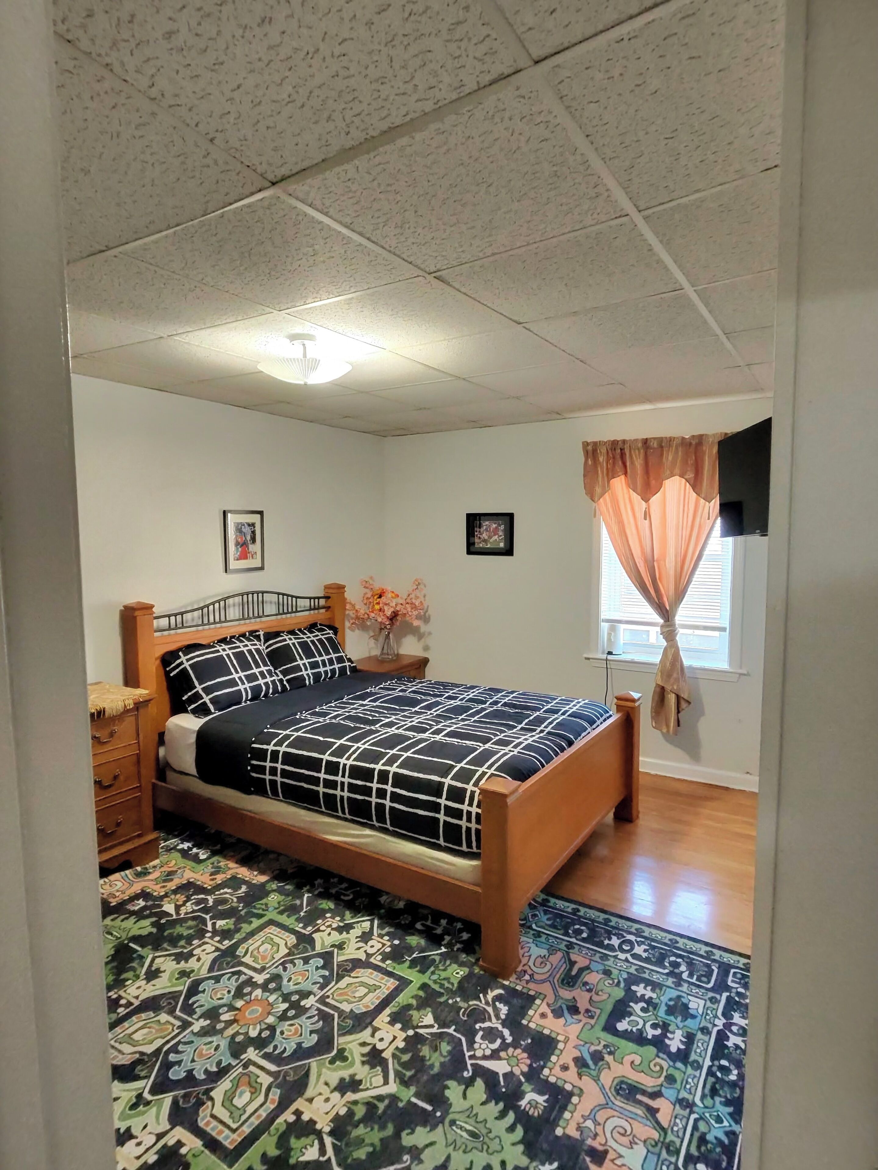 Un dormitorio con cama de madera, una mesita de noche, alfombra estampada, ventana con cortinas y una lámpara en el techo.