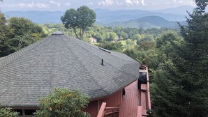 Une maison avec un toit en pente, entourée d’une végétation luxuriante et avec des montagnes en arrière-plan.