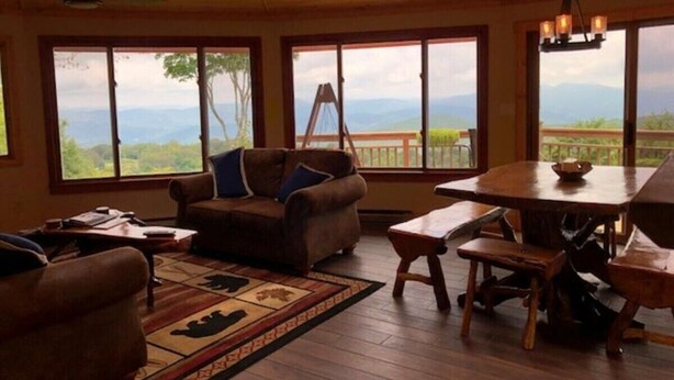 Un salon chaleureux avec un plafond en bois, de grandes fenêtres offrant une vue sur les montagnes, un canapé, une table à manger et un ventilateur de plafond.