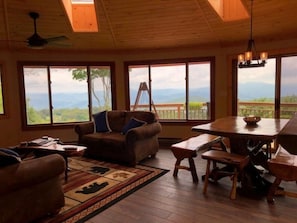 Ein gemütliches Wohnzimmer mit Holzbalkendecke, großen Fenstern mit Blick auf die Berge, einem Sofa, einem Esstisch und einem Deckenventilator.