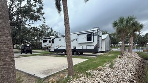 Un camping con dos vehículos recreativos estacionados en un área de grava, rodeado de árboles y un pequeño cuerpo de agua.