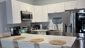 Eine moderne Küche mit weißen Schränken, einem Edelstahl-Kühlschrank, einer Mikrowelle und einer Kaffeemaschine. Um die Kochinsel stehen vier Stühle mit gewebten Platzsets.