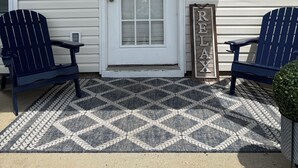 Eine Veranda mit einer weißen Tür, zwei Adirondack-Stühlen, einem gemusterten Teppich und Wanddekoration, darunter ein Strandkranz und Fische.