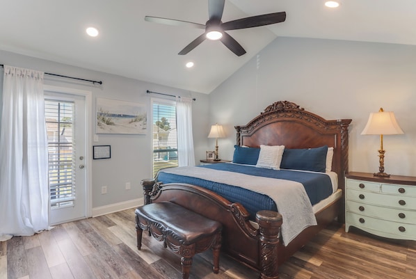 A bedroom with a large bed, a ceiling fan, a bench, a nightstand, and a window with blinds.