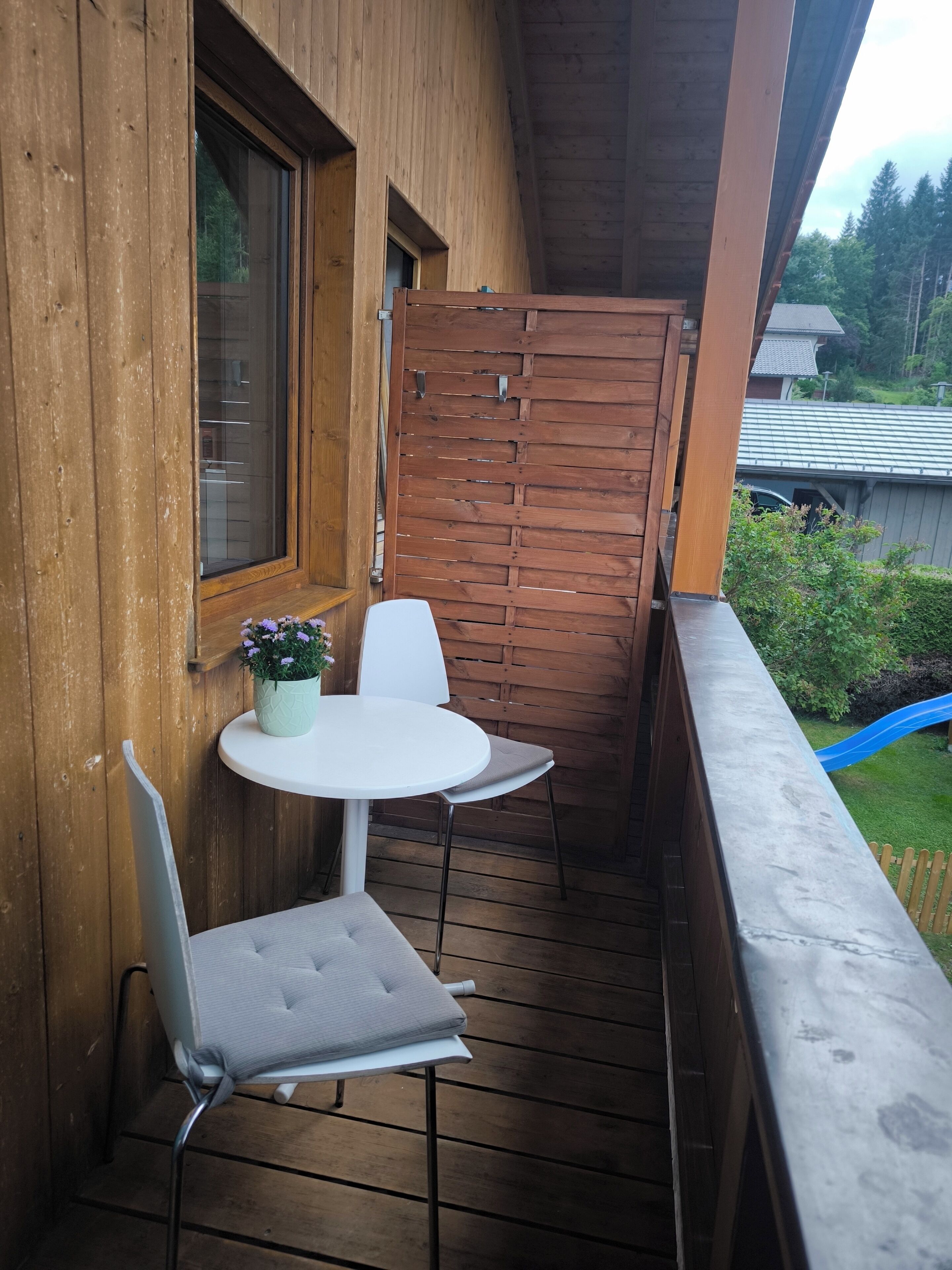 A wooden balcony with a white table and chairs, a potted plant, and a view of a blue slide and greenery.