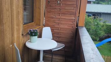 A wooden balcony with a white table and chairs, a potted plant, and a view of a blue slide and greenery.