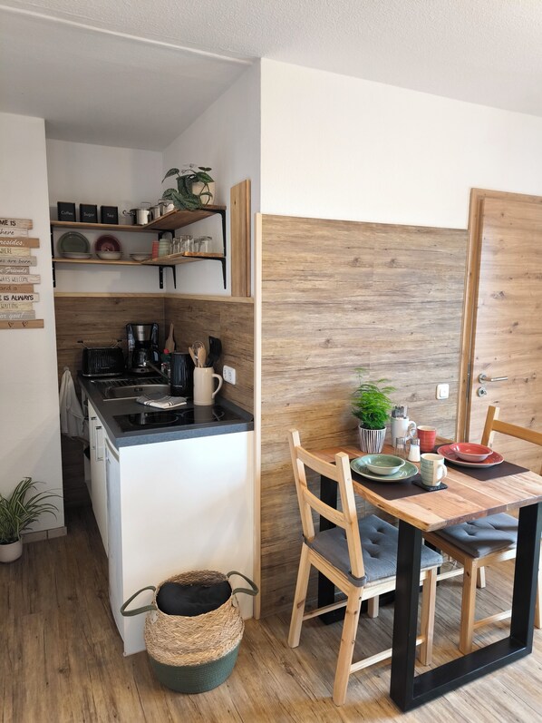 A compact kitchen with a stove, coffee maker, and shelves above. A dining table with chairs and a small plant is visible.