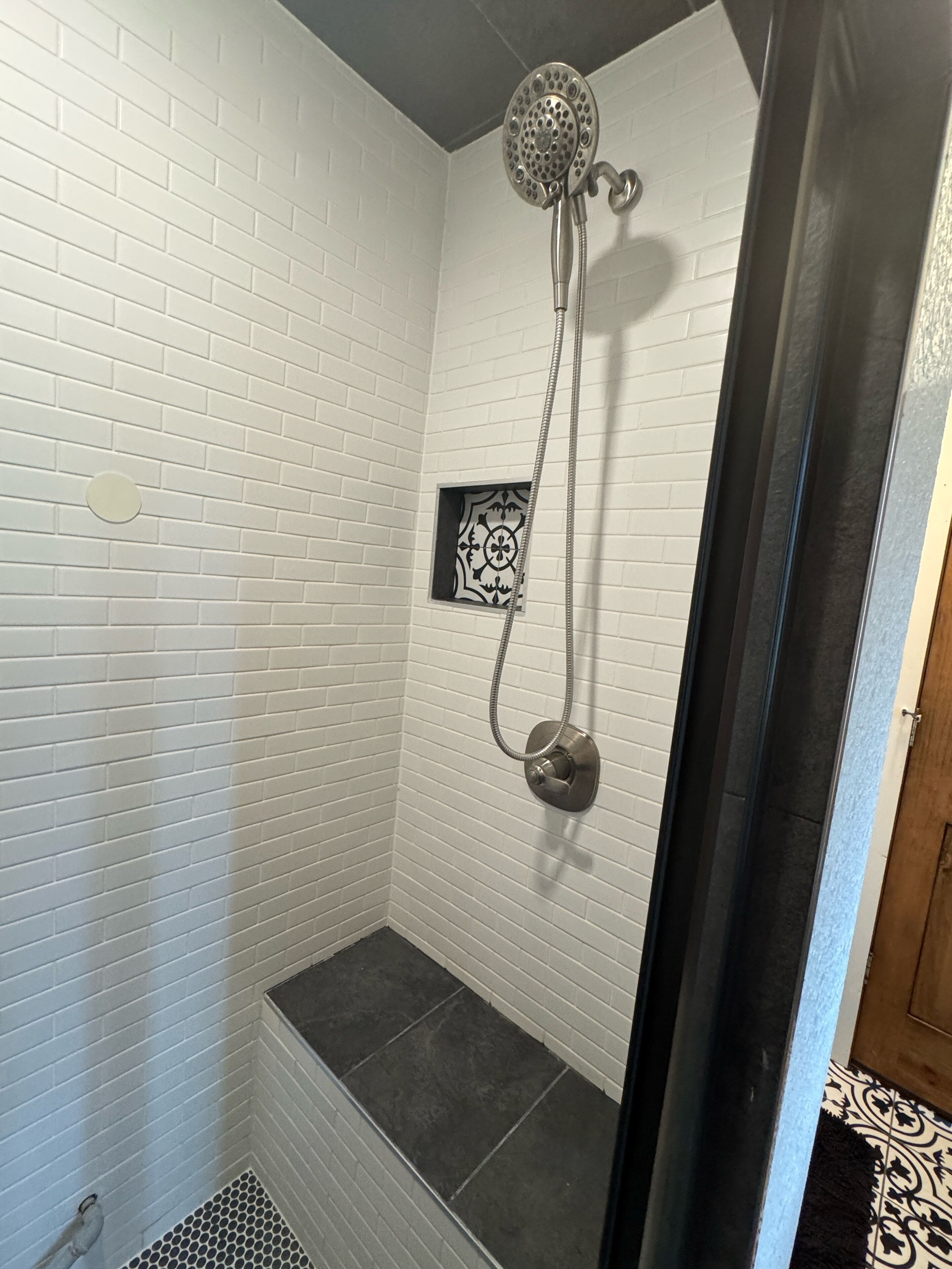 Une douche équipée d’un pommeau de douche à main, avec des murs recouverts de carreaux blancs et une petite fenêtre décorative.