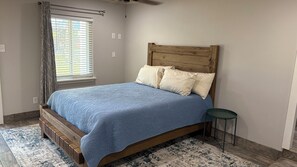 A bedroom with a wooden bed frame, a blue bedspread, two pillows, a small round table, and a window with blinds. - Lake Cottage 200 yards from the Lake Texoma shoreline! (Pottsboro)