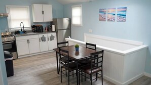 Una cocina compacta con armarios blancos, un refrigerador de acero inoxidable y una estufa a gas. Hay una zona de comedor con una mesa y sillas de madera, y una pequeña planta en maceta sobre la mesa.