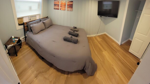 Un dormitorio con una cama grande, una mesita de noche, un televisor montado en la pared y una ventana con persianas.