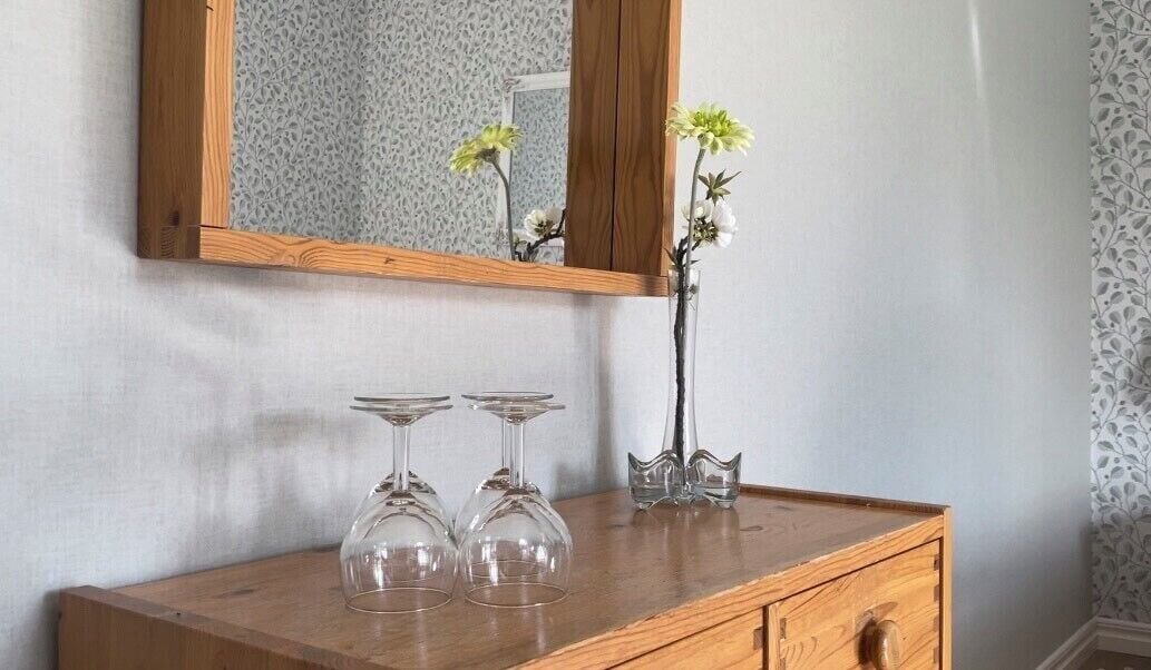 Une commode en bois avec un miroir au-dessus, un vase rempli de fleurs et deux verres à vin posés sur la commode.