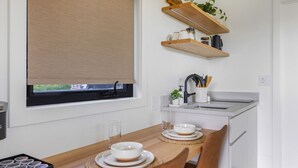 Een compacte keuken met een eethoek, met een raam voorzien van een beige zonwering, houten planken boven het aanrecht en een kleine potplant.