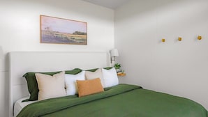 Una cama bien hecha con una colcha verde y varios cojines, una mesita de noche con una lámpara y un cuadro con un paisaje en la pared.