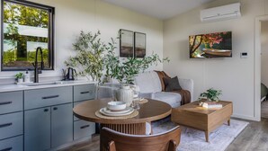 Una cocina moderna con gabinetes grises, una mesa de comedor redonda y una silla de madera. Hay un sofá y una mesita de centro con libros y una planta.