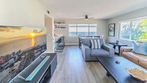 Ein modernes Wohnzimmer mit einem großen Fernseher, der einen Sonnenuntergang zeigt, einem Essbereich mit Tisch und Stühlen, einer Couch mit Kissen und einem Fenster mit Blick auf Grünflächen.